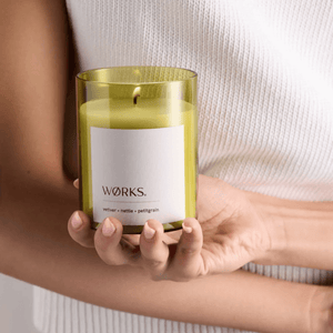 Hand-poured WØRKS candle in a simple glass jar, surrounded by natural elements like green leaves and herbs, evoking a calm and balanced atmosphere.