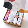 A luxurious selection of self-care products including a hydrating body cream, organic chai tea, soy candle, sparkling botanical drink, lavender-filled eye pillow, and a natural soap bar – all beautifully arranged to create a relaxing spa experience at hom