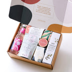 A luxurious selection of self-care products including a hydrating body cream, organic chai tea, soy candle, sparkling botanical drink, lavender-filled eye pillow, and a natural soap bar – all beautifully arranged to create a relaxing spa experience at hom