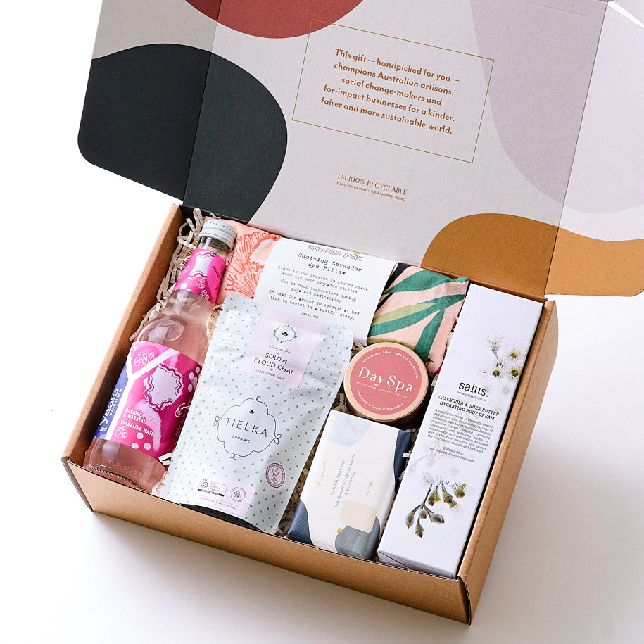 A luxurious selection of self-care products including a hydrating body cream, organic chai tea, soy candle, sparkling botanical drink, lavender-filled eye pillow, and a natural soap bar – all beautifully arranged to create a relaxing spa experience at hom
