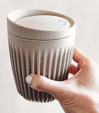 Hand holding a textured ceramic cup with a lid against a light background included in the hamper of curated with conscience