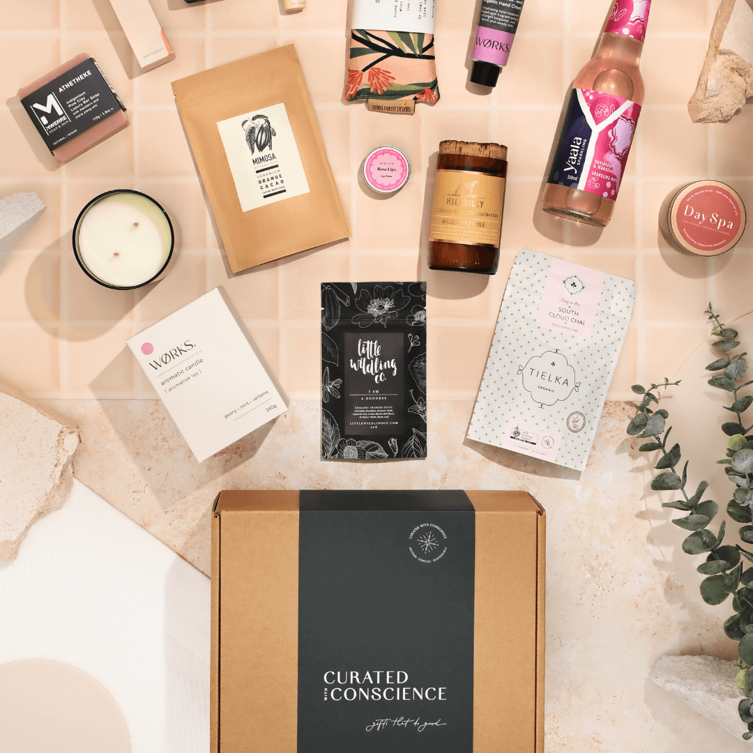 A beautifully arranged vegan hamper featuring eco-friendly gifts including candles, skincare products, gourmet snacks, artisan chocolate, tea, and self-care essentials from Curated with Conscience, presented in a sustainable kraft box on a neutral tiled background
